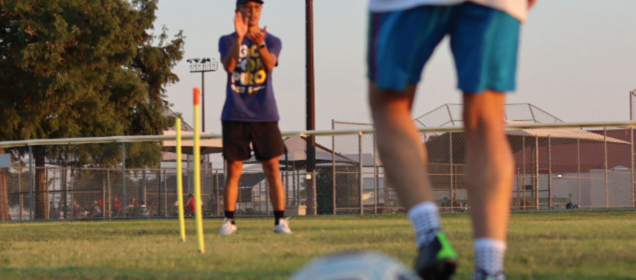 Adult athletes in open-play soccer game with improper form, USSPT coach preparing to guide skills training.